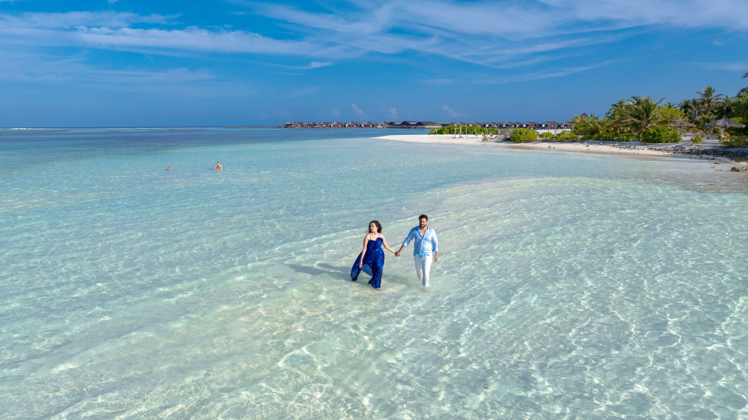 A couple walking along the clear waters of a Maldives beach, under a vibrant blue sky.